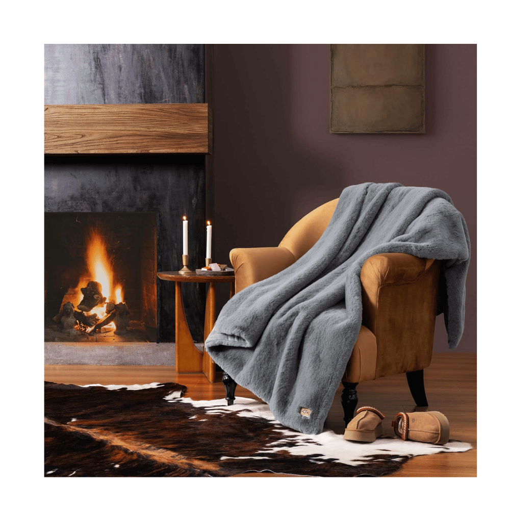 Gray UGG Plush Faux Fur Throw Blanket draped over armchair by fireplace, creating a cozy home setting.