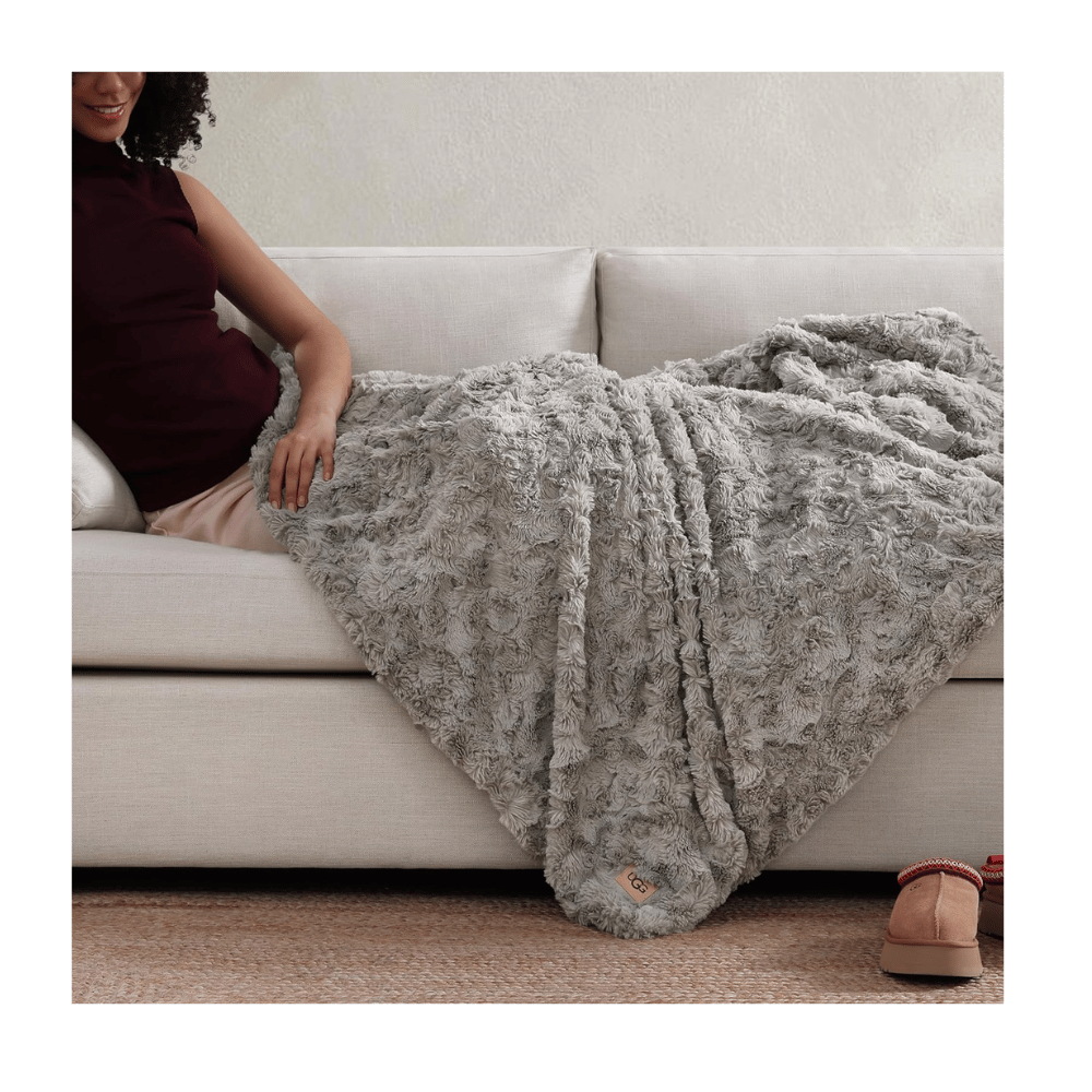 Woman sitting on sofa covered with UGG Adalee Faux Fur Throw Blanket in grey faux fur (Credit: MyRadiantSleep.com)